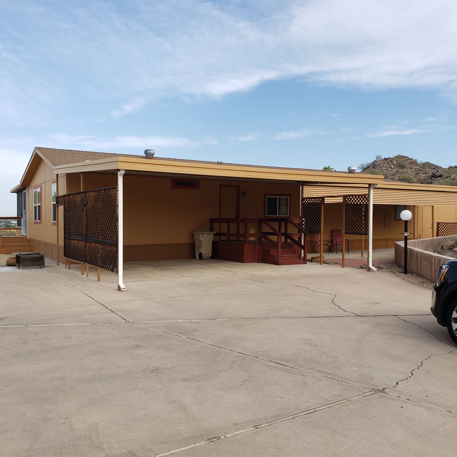 Tan manufactured home with carport. Concrete driveway in desert setting.