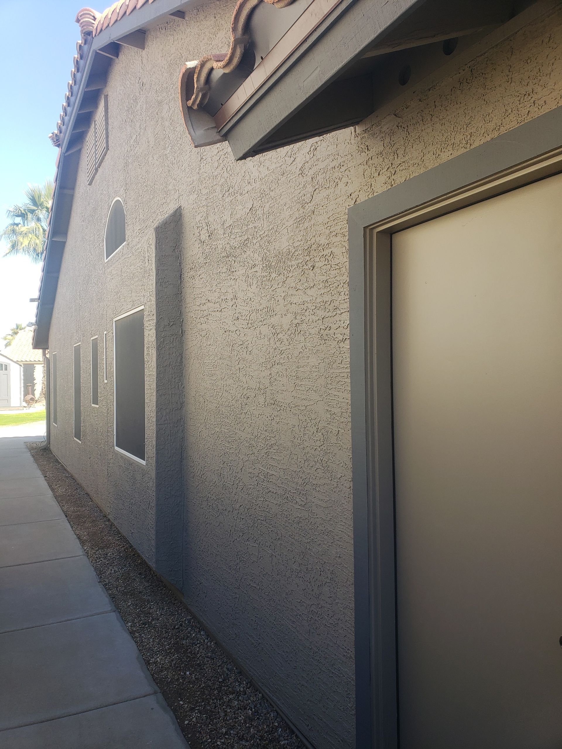 Exterior of a stucco building with a doorway and windows, next to a walkway with a rock bed, under a blue sky.
