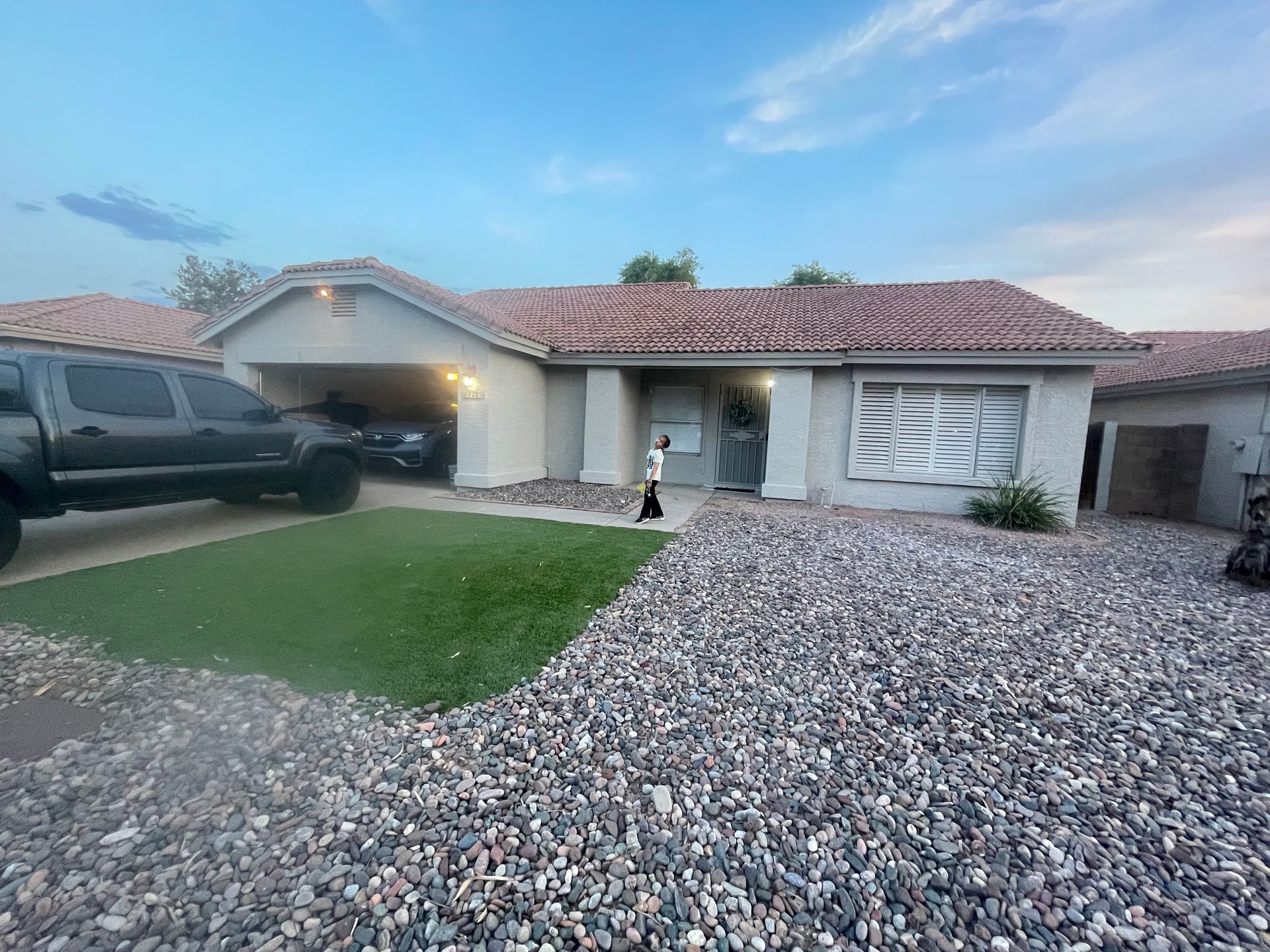Gray truck parked next to a beige house with a gravel yard and green turf; sky is blue with some clouds.