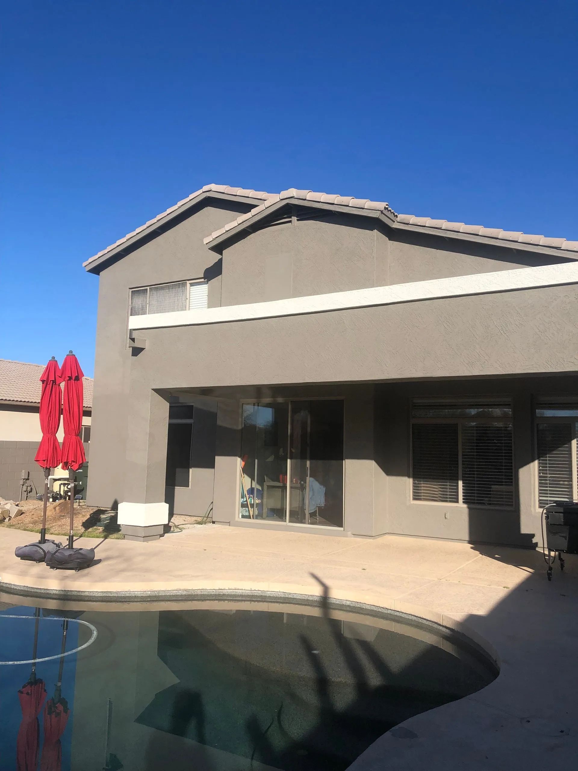 Backyard with pool, gray stucco house, red umbrella, patio furniture, and clear blue sky.