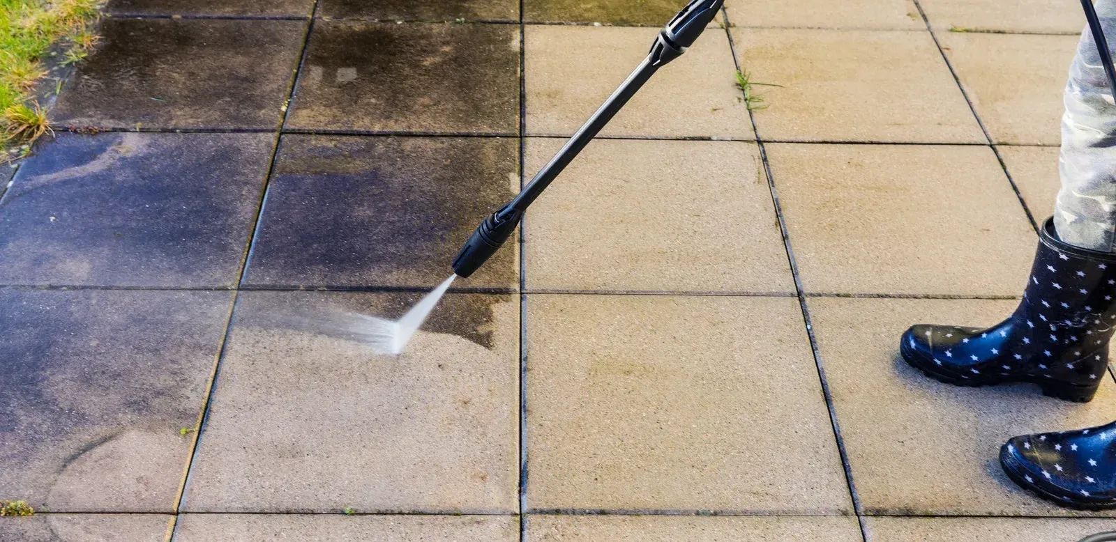 Pressure washing dirty stone patio beside a person’s boots, with a spray wand cleaning dark stains.