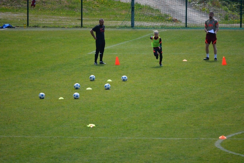 Soccer training on a grassy field. A child runs toward cones and soccer balls, with two coaches observing.