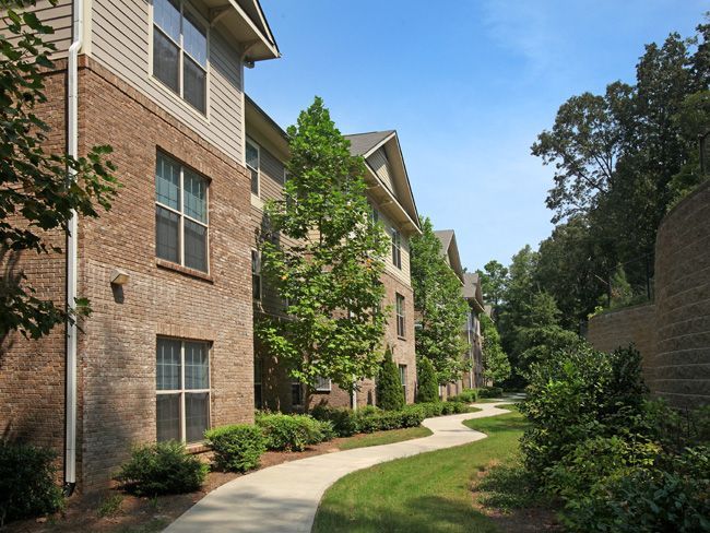 Brick apartment buildings with a winding sidewalk, surrounded by green trees and bushes under a blue sky.