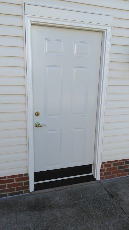 A white, six-panel door set in light-colored siding above a brick foundation and concrete patio.