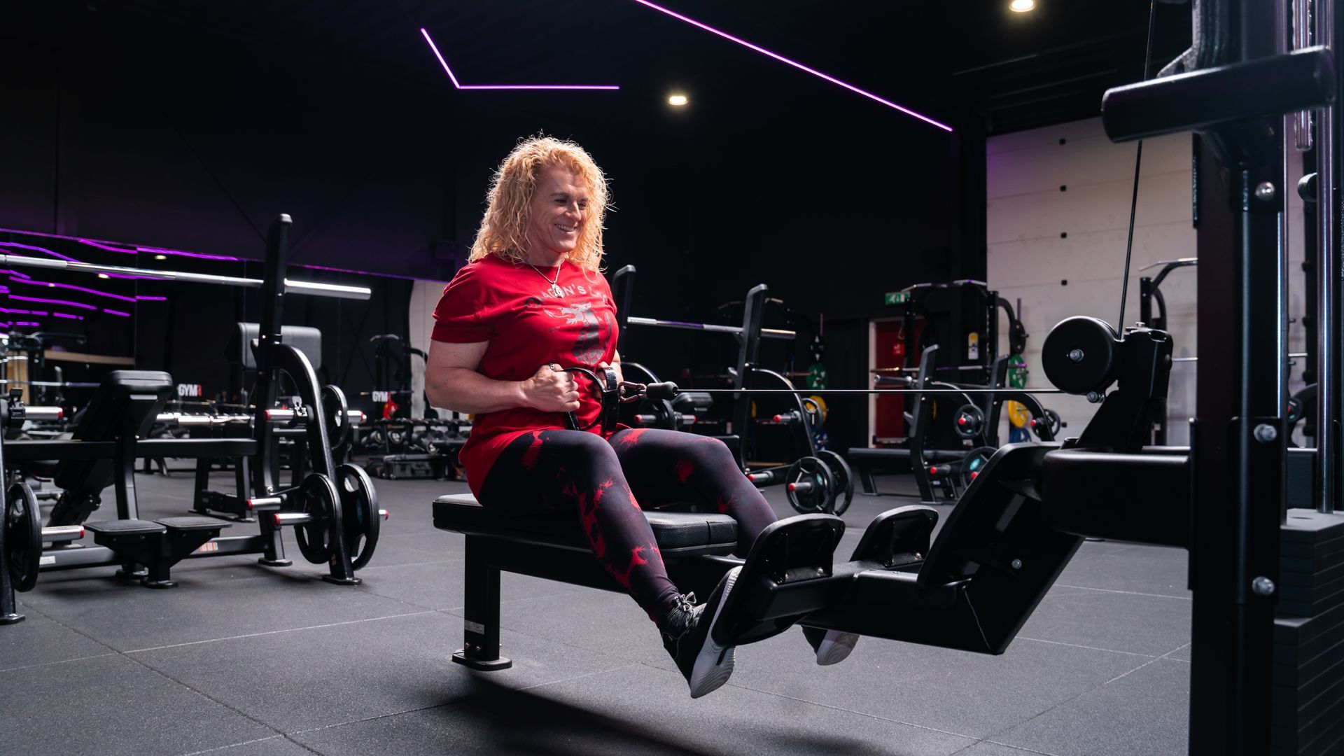 Man in de sportschool doet lat pulldowns; zwart tanktopje, donkere setting met paarse neonaccenten.