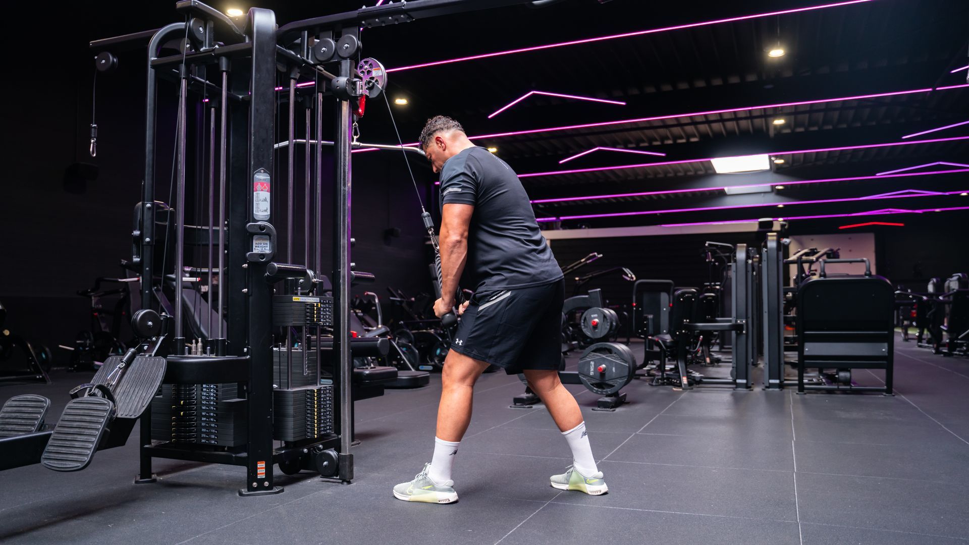 Een persoon tilt een halter boven zijn hoofd terwijl hij op een schuine bank ligt in een sportschool met paarse neonverlichting.