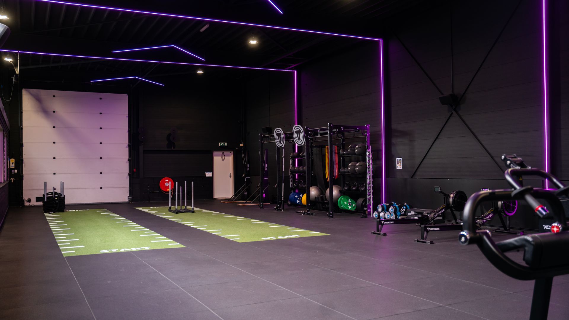 Interieur van de sportschool met apparatuur en een groene baan op de vloer; donkere verlichting en verschillende mensen die aan het trainen zijn.