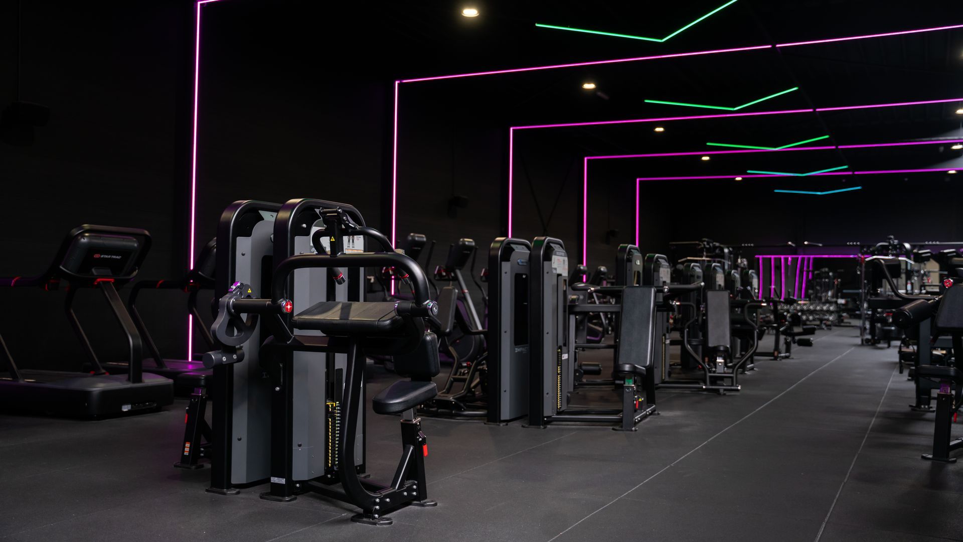 Donkere sportschool met fitnessapparaten, paarse plafondlampen en een witte muur.