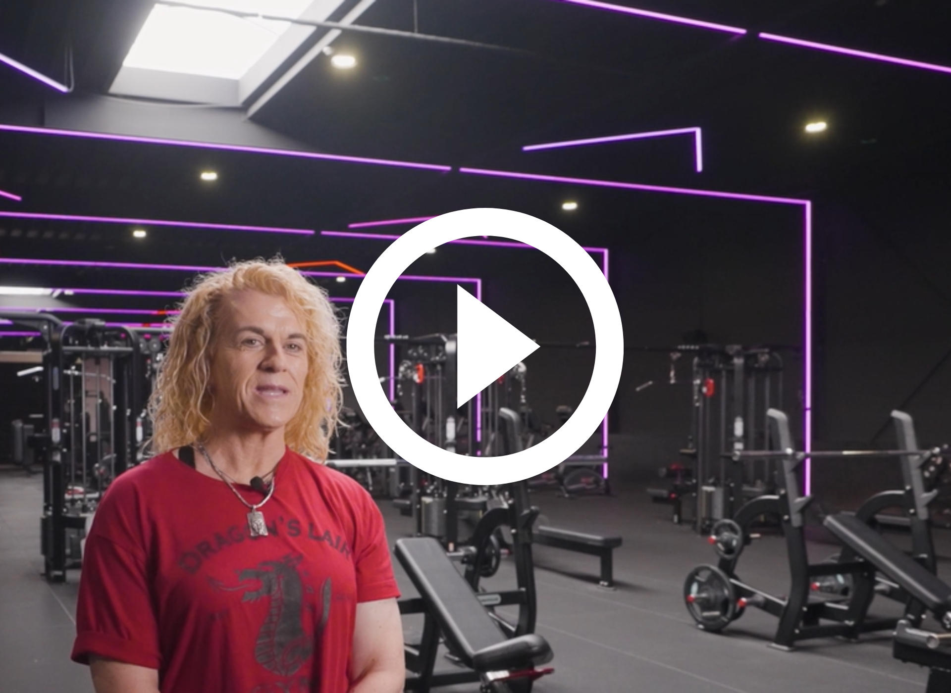 Man in een sportschool; praat in de camera; afspeelknop eroverheen; sportschoolomgeving met apparatuur en mensen op de achtergrond.