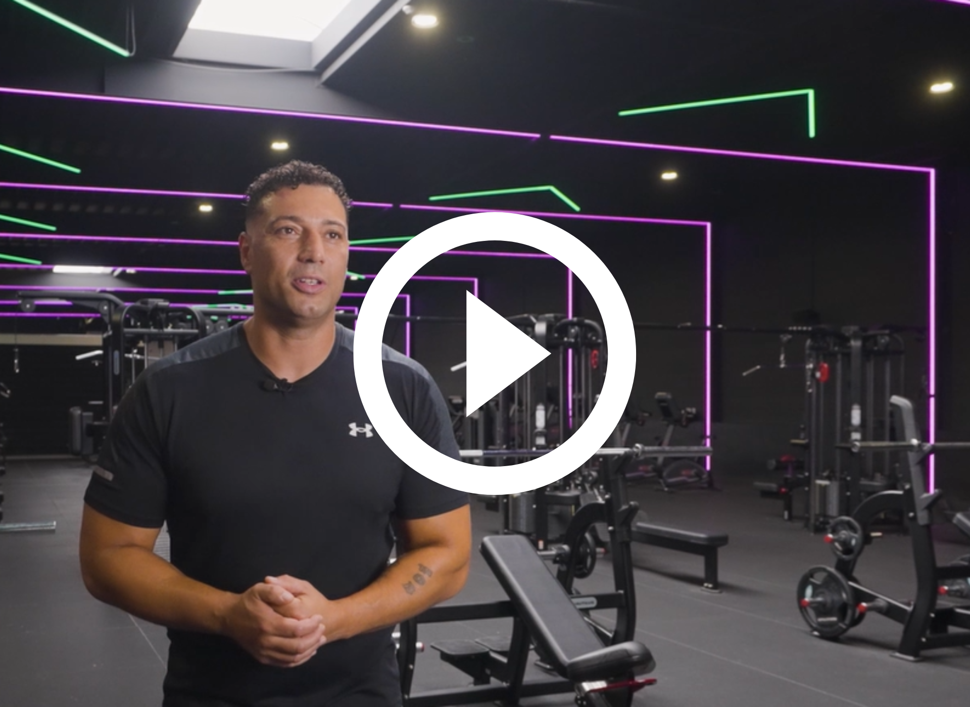 Vrouw in de sportschool, gekleed in een zwart tanktopje, lachend. Fitnessapparatuur en mensen op de achtergrond. Afspeelknop erboven.