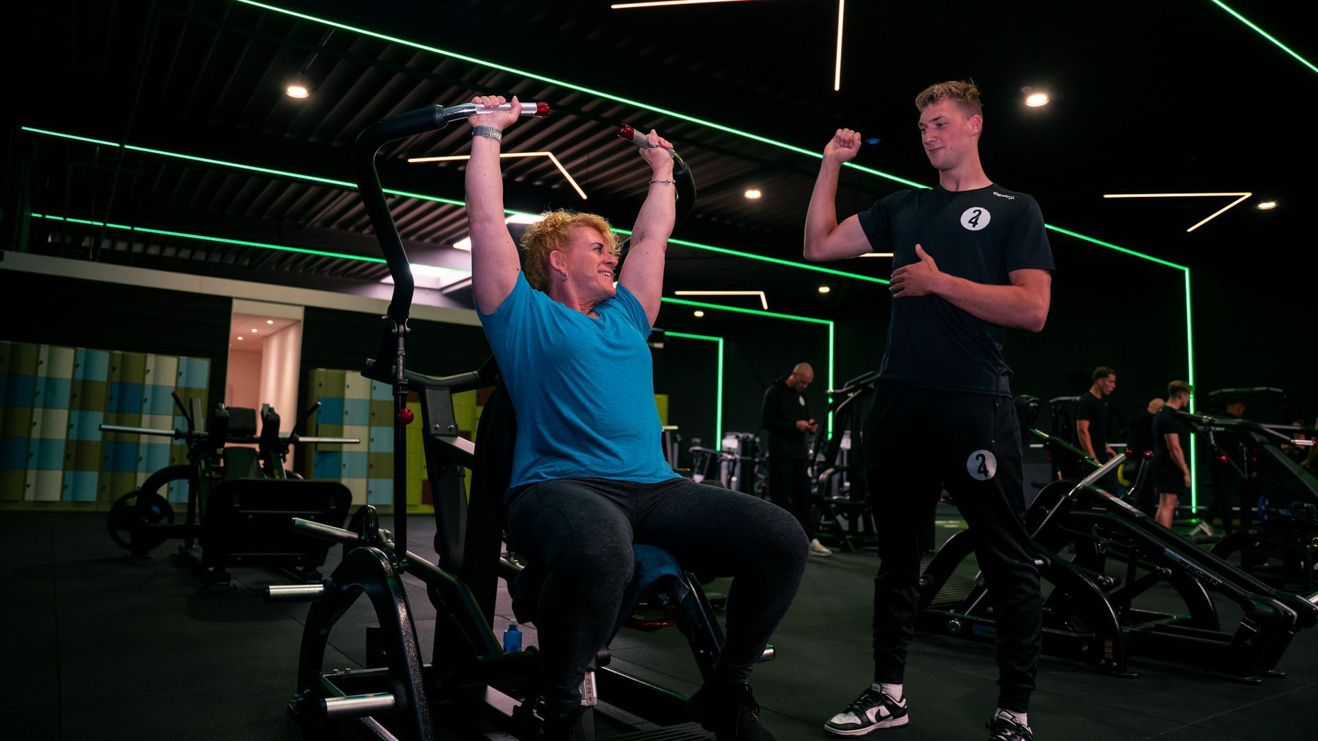 Vrouw traint met een trainer in de sportschool, tilt gewichten boven haar hoofd op een apparaat, met groene neonaccenten.