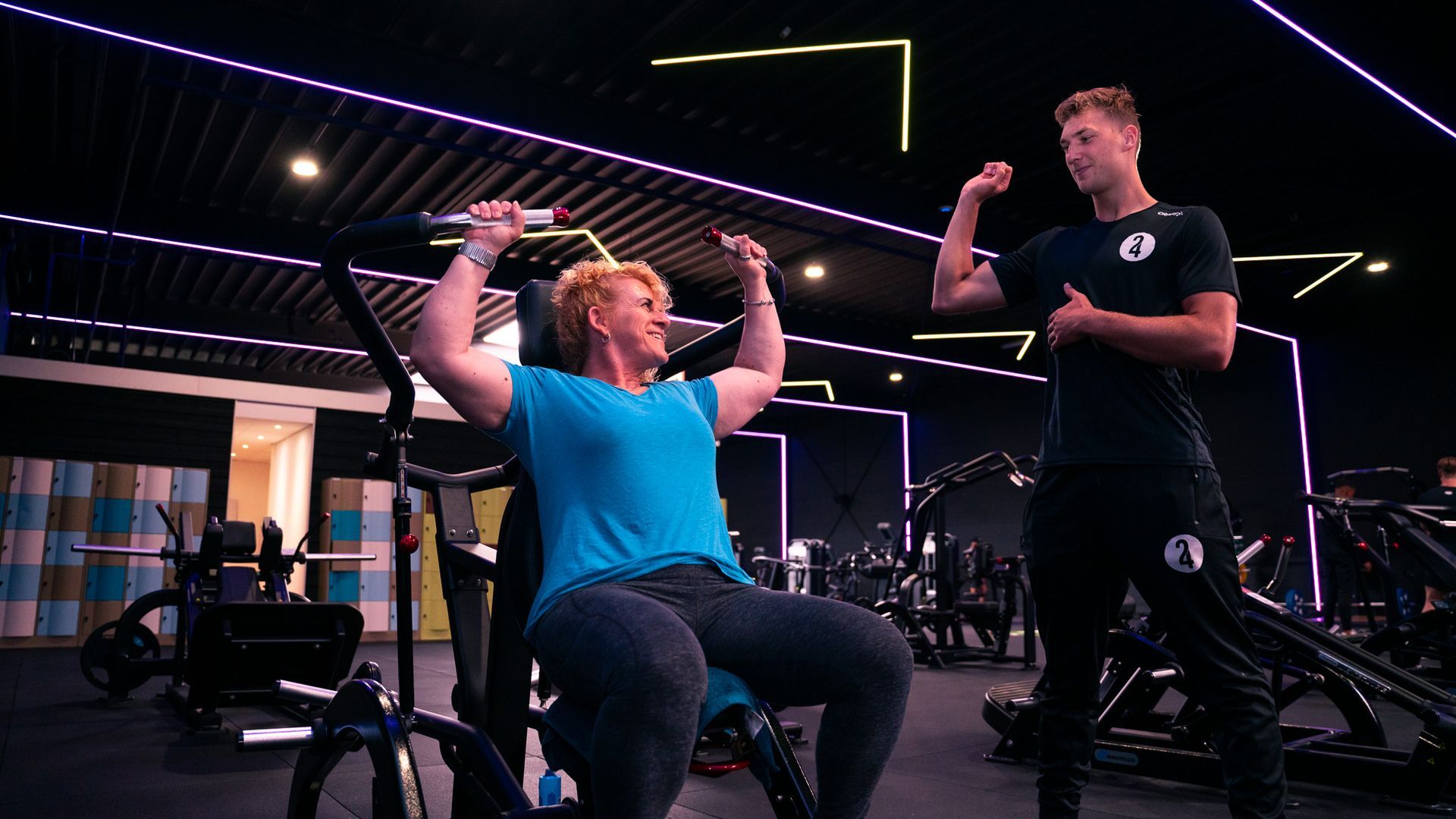 Een vrouw is in de sportschool aan het trainen met een trainer. Zij gebruikt een apparaat om gewichten te tillen en hij spant zijn biceps aan.