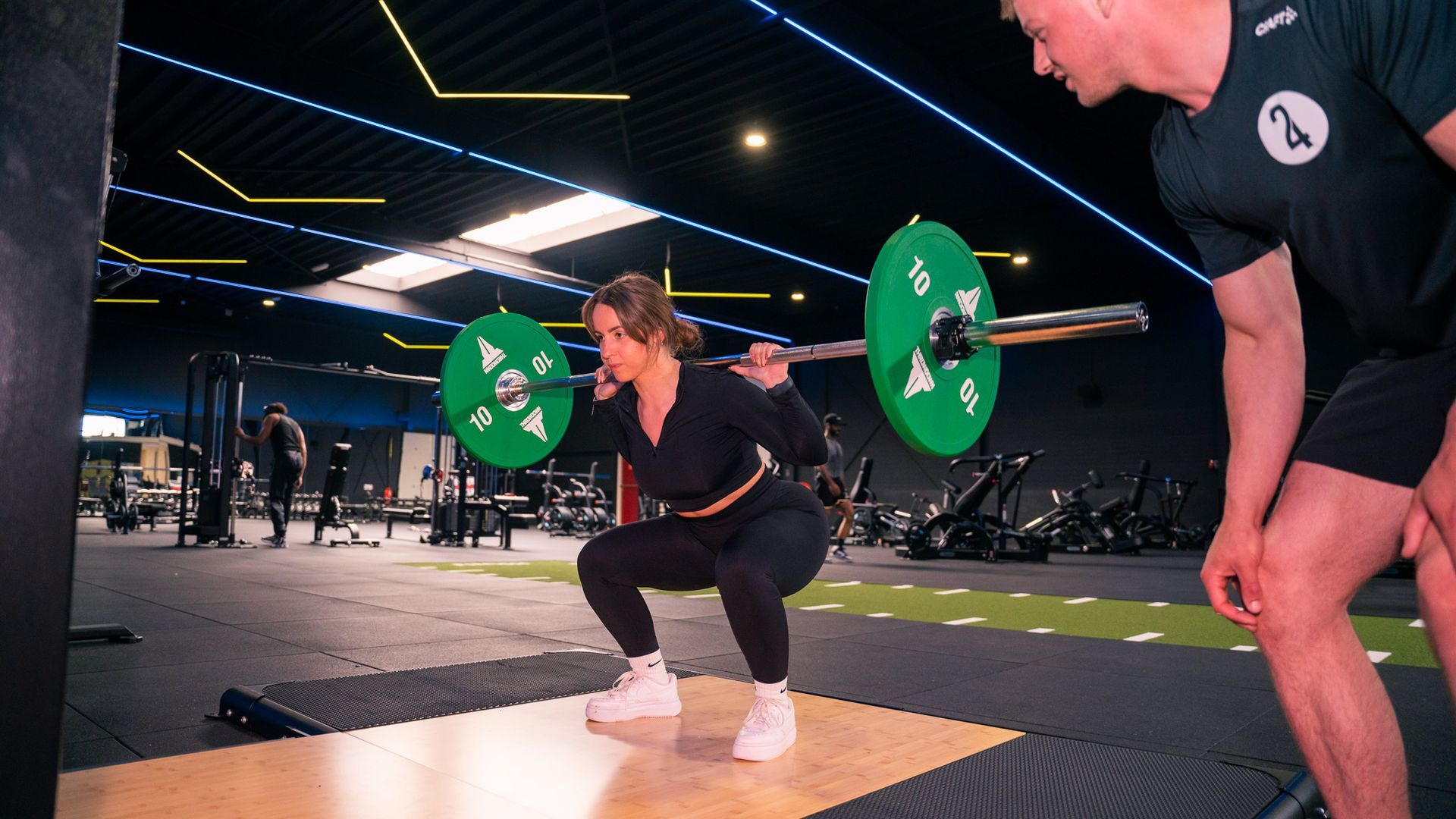 Vrouw squat met halterstang, begeleid door een trainer, in een sportschool. Groene gewichten aan de stang. Zwarte sportkleding.