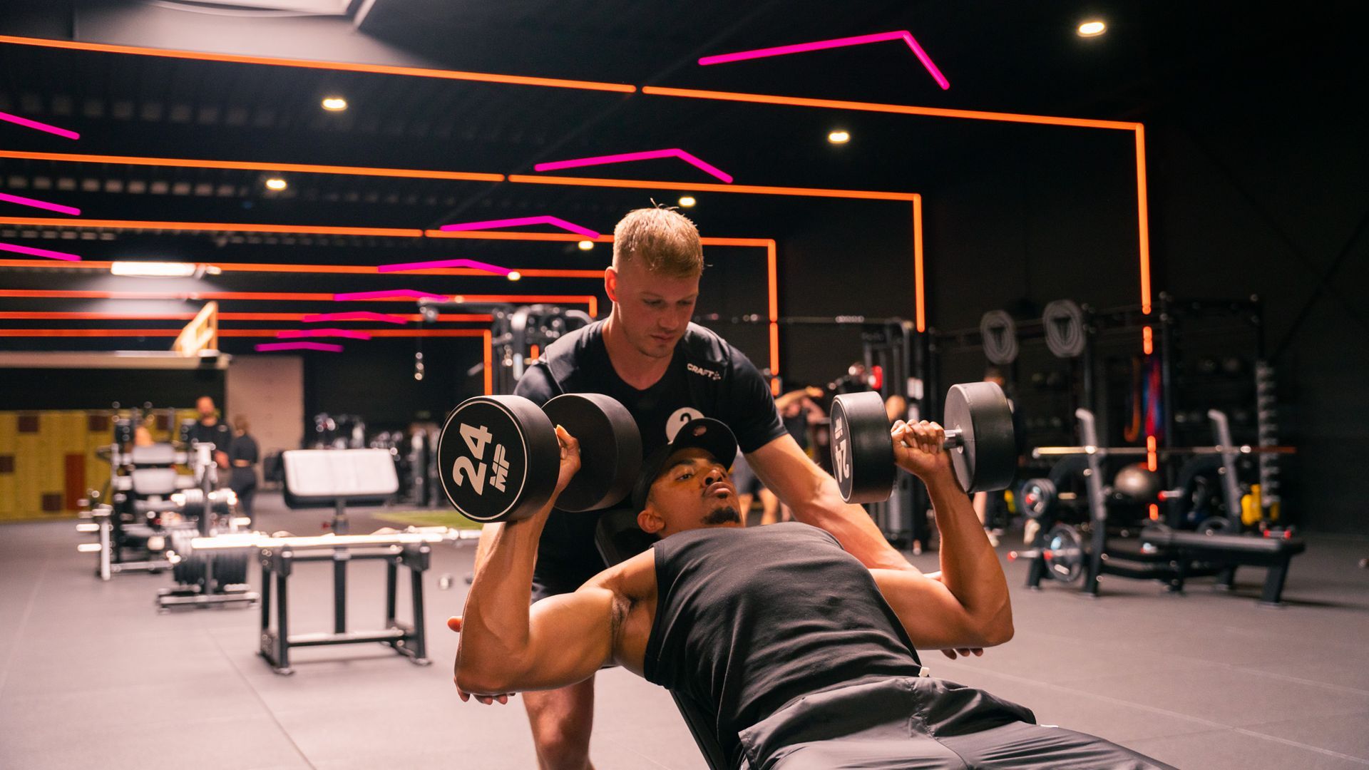 Man is aan het bankdrukken met een trainer in de gaten in een neonverlichte sportschool.