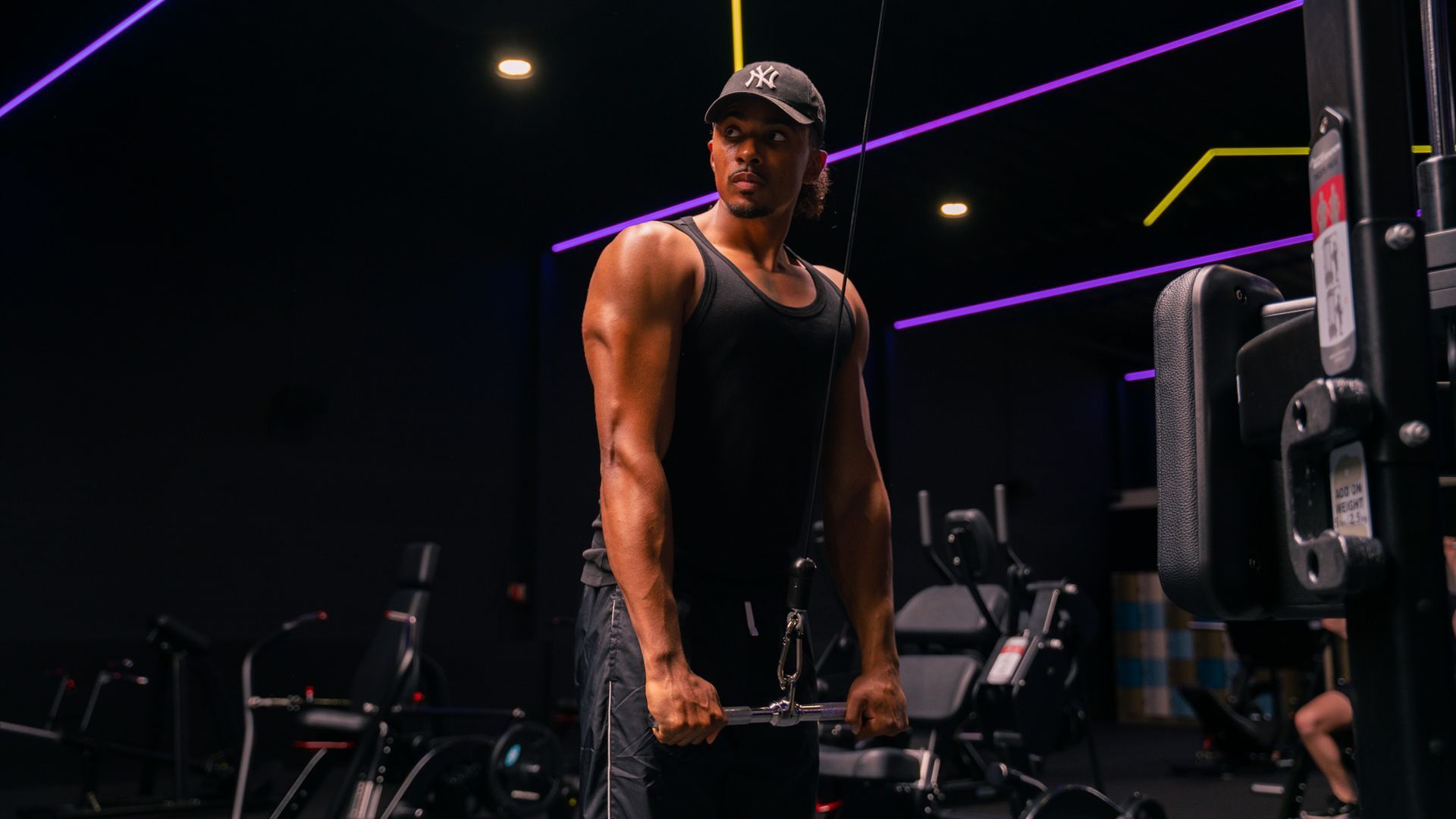 Man in een zwart tanktopje in de sportschool, doet tricep cable pushdowns.