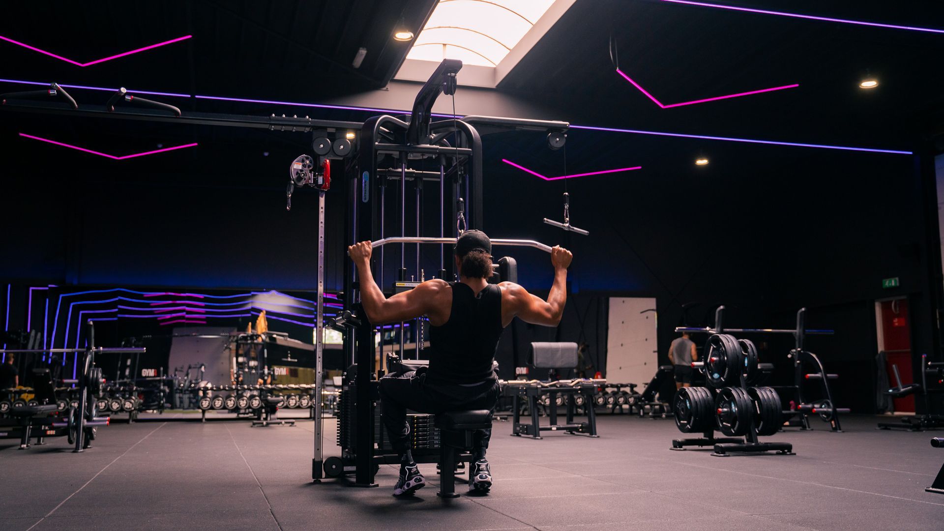 Man doet een lat pulldown in de sportschool. Hij draagt zwart en staat met zijn rug naar de camera. Donkere kamer met fitnessapparatuur.