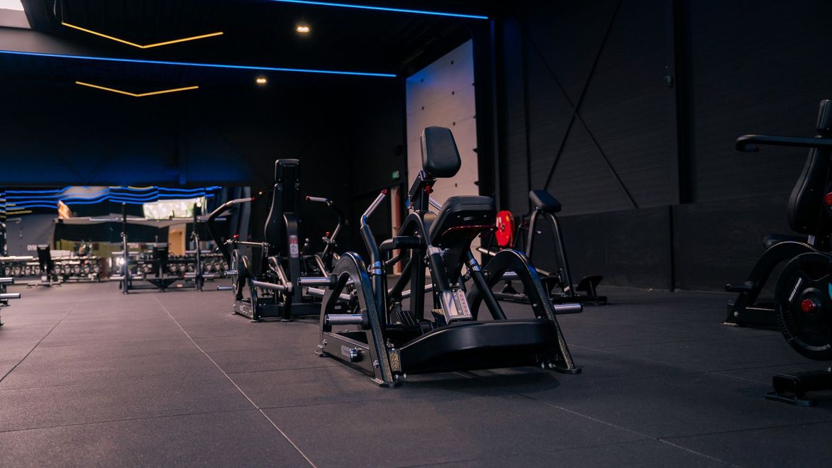 Interieur van een sportschool met fitnessapparaten, donkere muren en blauwe accentverlichting.