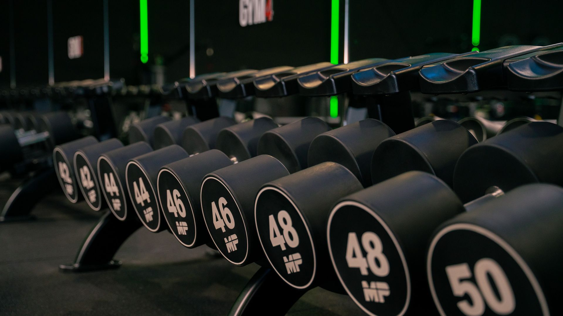 Halters in een sportschool, gerangschikt op gewicht. Zwarte gewichten op zwarte rekken. Genummerde labels: 44, 46, 48, 50.