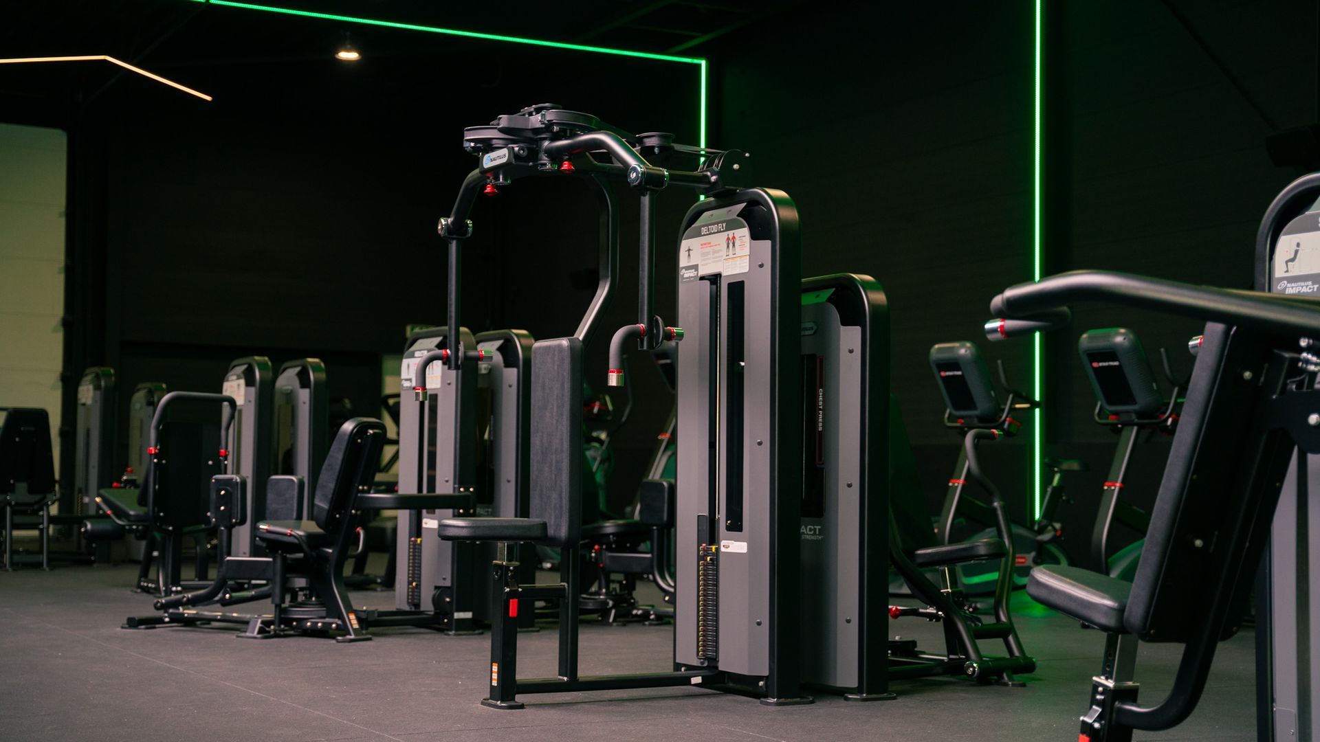Interieur van een sportschool met donkere muren, fitnessapparaten en groene accentverlichting.