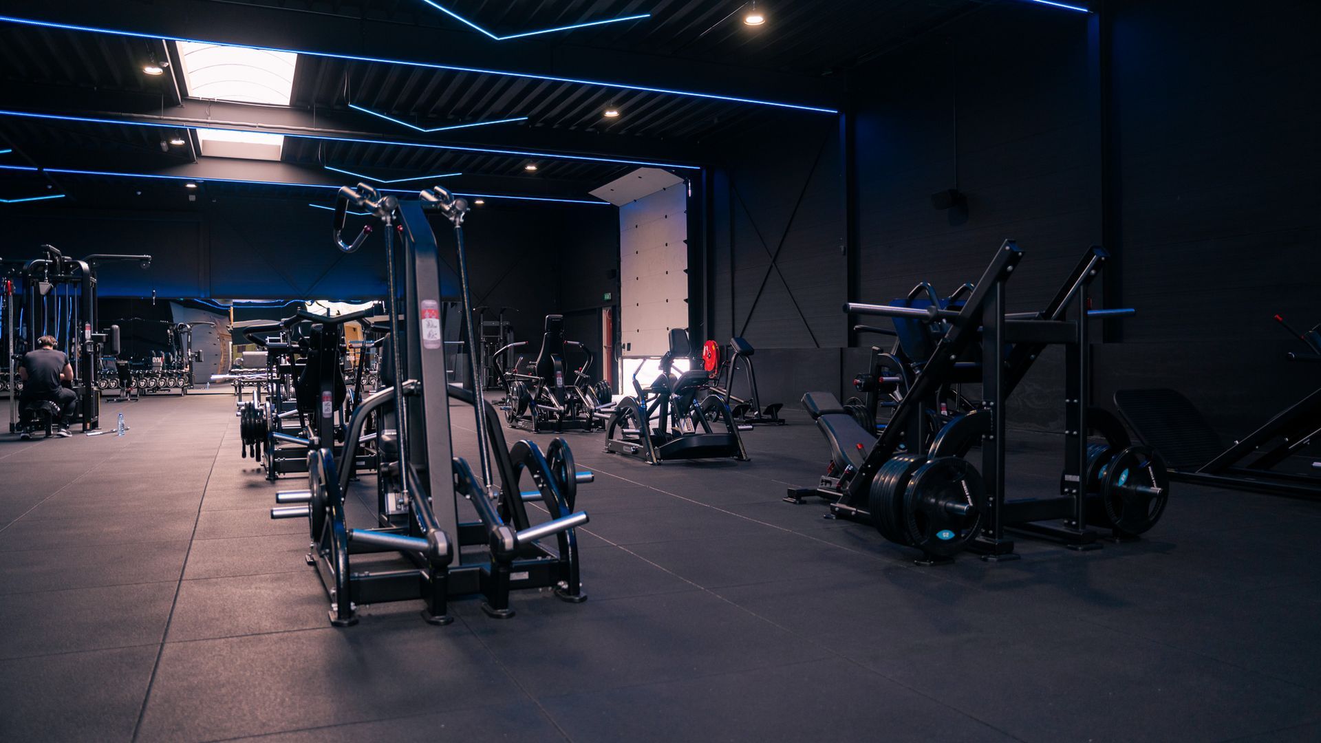 Interieur van een sportschool met fitnessapparaten op een zwarte vloer, blauwe accentverlichting en donkere muren.