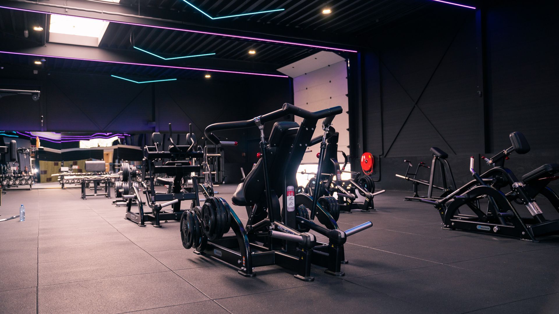 Donkere sportschool met fitnessapparaten, paarse plafondlampen en een witte muur.
