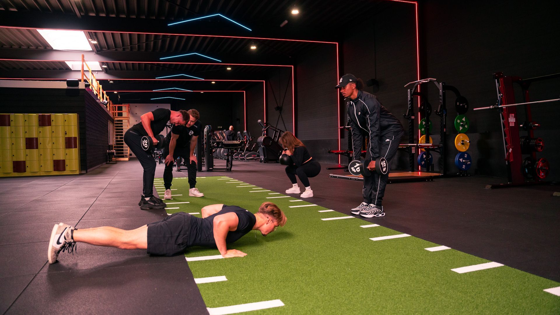 Mensen trainen in een sportschool. De een doet push-ups, de ander tilt halters. Anderen kijken toe. Groene grasmat.