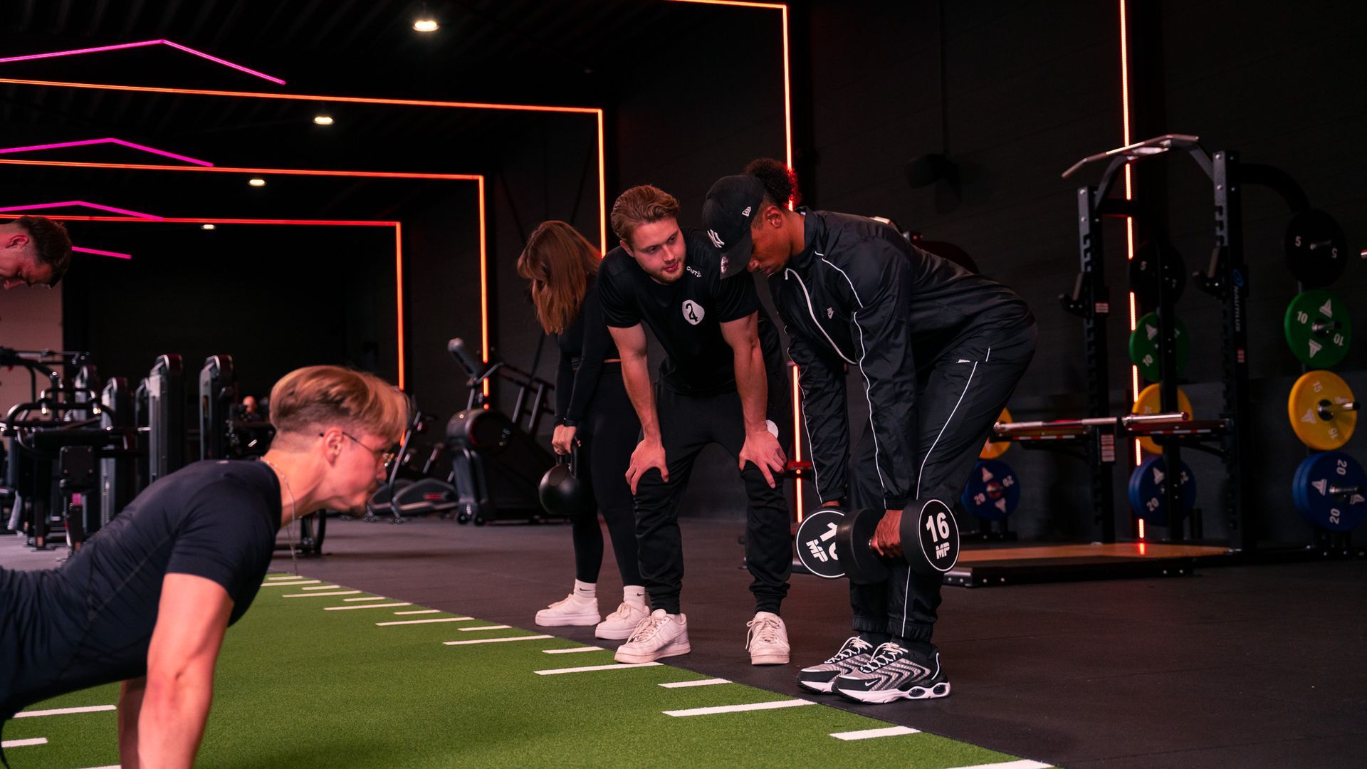 Mensen in een sportschool: een man met gewichten en drie anderen kijken toe en helpen hem.