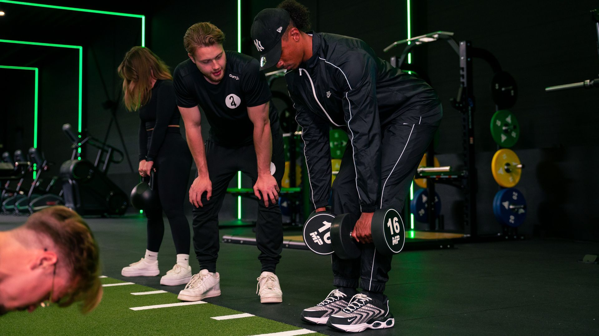 Sportsessie: Trainer begeleidt iemand die halters tilt. Twee mensen kijken toe. Groene sportschool.