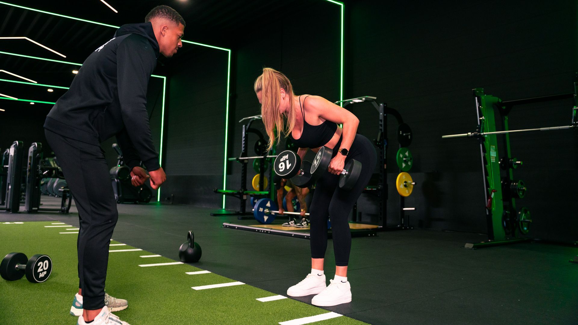 Een vrouw doet in de sportschool een bent-over row met halters, onder begeleiding van een man.