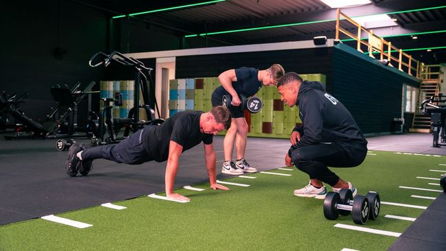 Drie mannen in een sportschool: één doet push-ups, één met een halter en één kijkt toe.