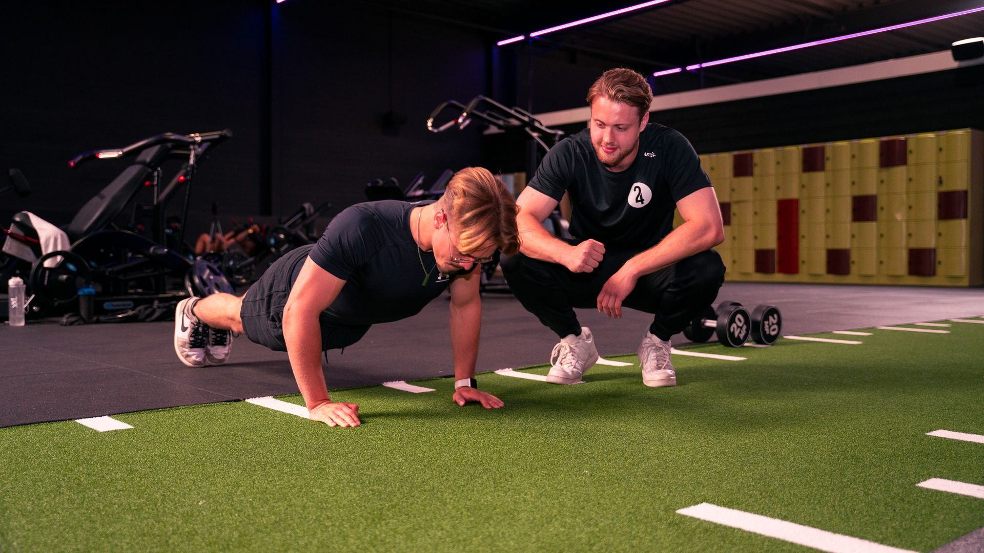 Een man doet push-ups op een groene ondergrond, terwijl een coach toekijkt. Sportschoolomgeving.