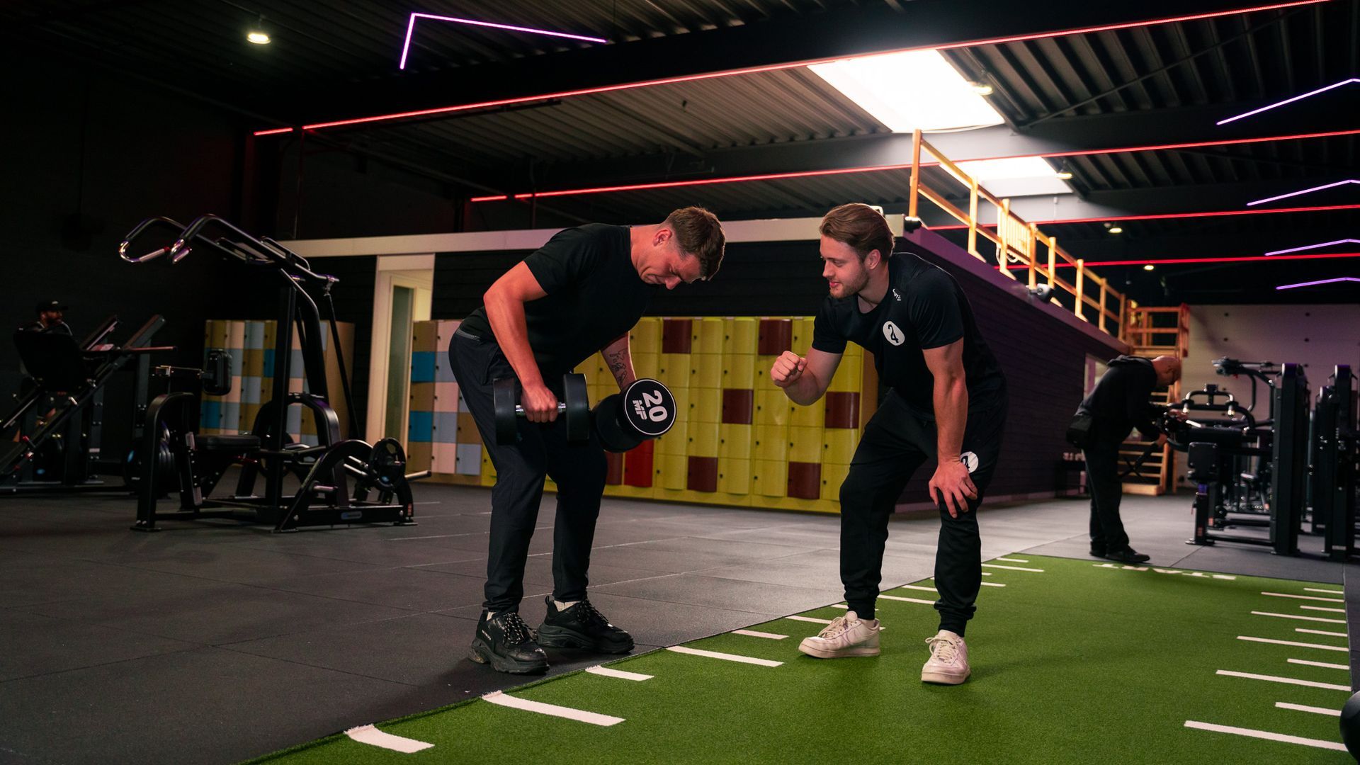 Twee mannen in een sportschool; de een tilt een halter terwijl de ander coacht.