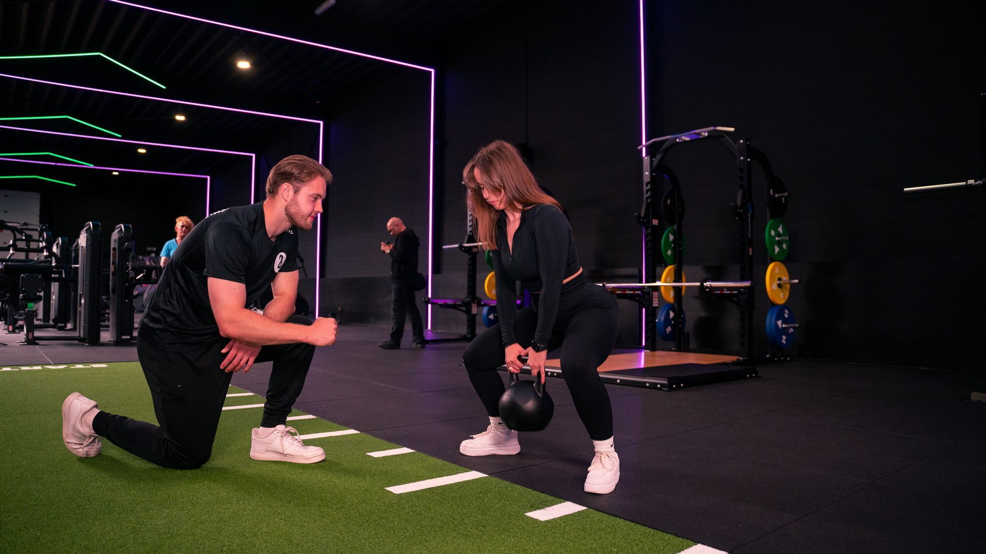 Een vrouw doet een kettlebell squat, gecoacht door een man, in een sportschool met neonverlichting.
