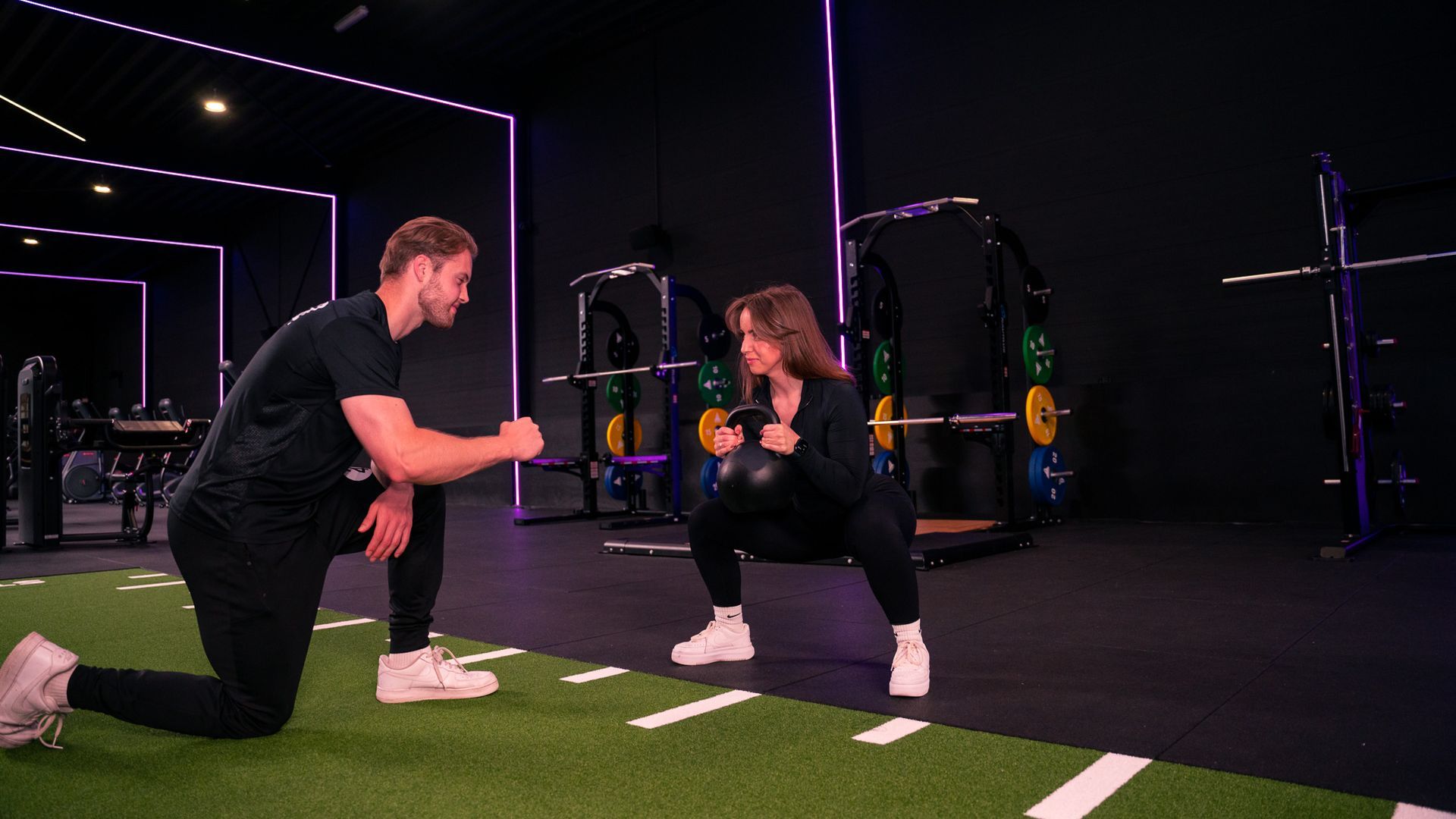 Een vrouw doet een squat met een gewicht terwijl een trainer toekijkt in een sportschool met kleurrijke verlichting.