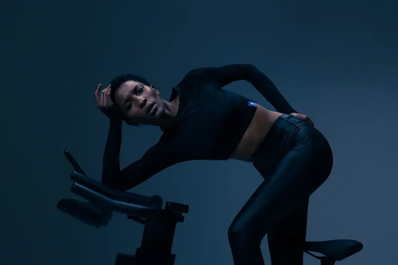 Een vrouw in zwarte sportkleding leunt op een hometrainer. Ze ziet er moe uit en heeft een blauwe achtergrond.