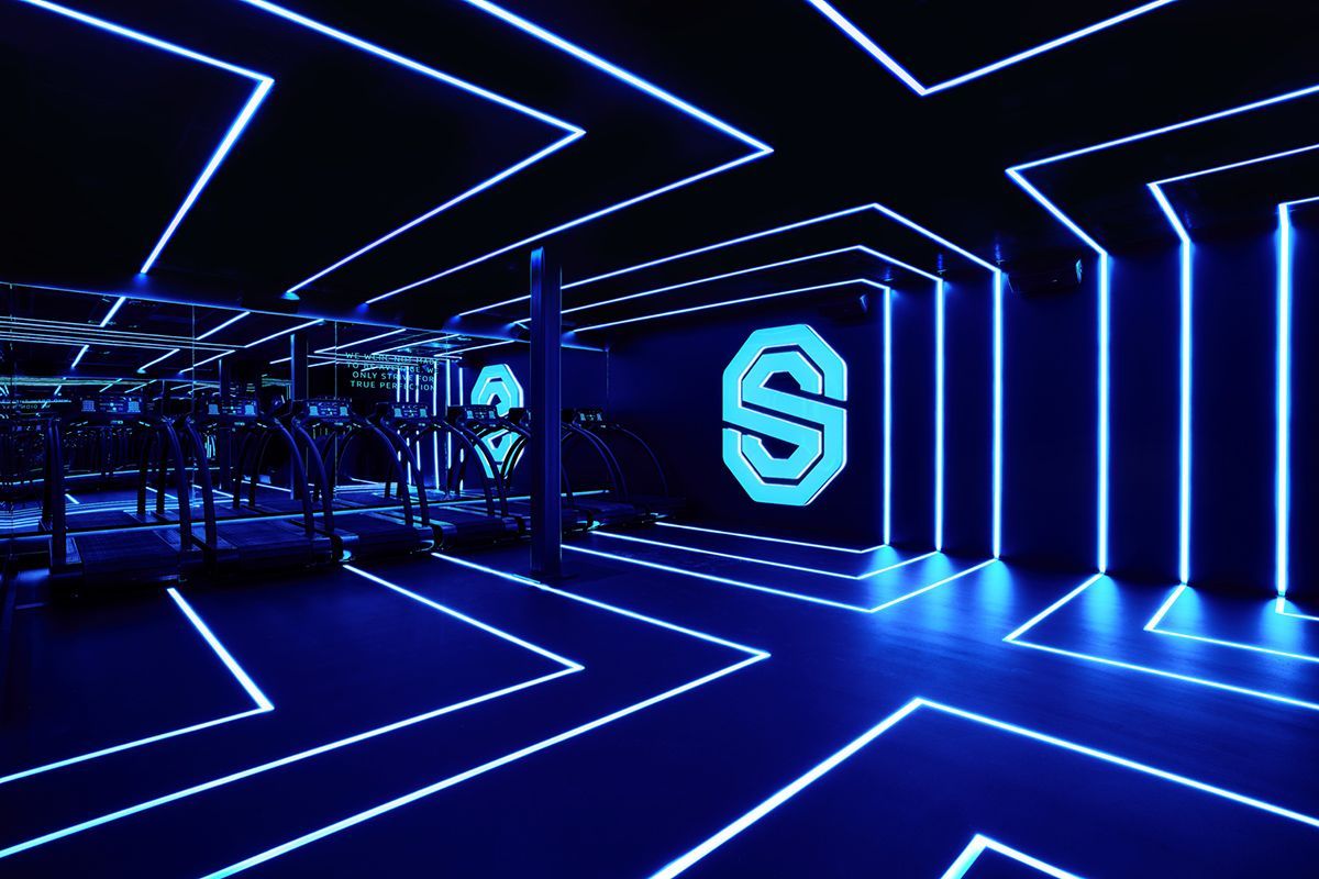 Blauwe neonverlichte sportschool met een logo, fitnessapparatuur en geometrisch plafondontwerp.