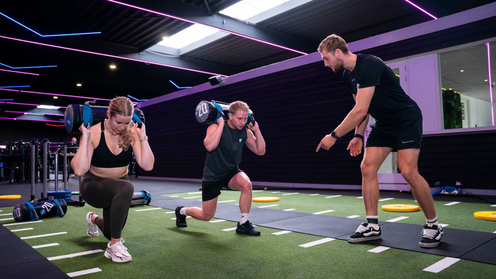 Vrouw doet lunges met halters, onder begeleiding van een coach, in een sportschool met neonverlichting.