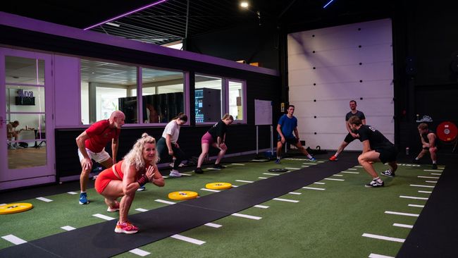 Drie mannen trainen in een sportschool. Eén doet push-ups terwijl de anderen toekijken en gewichten heffen.