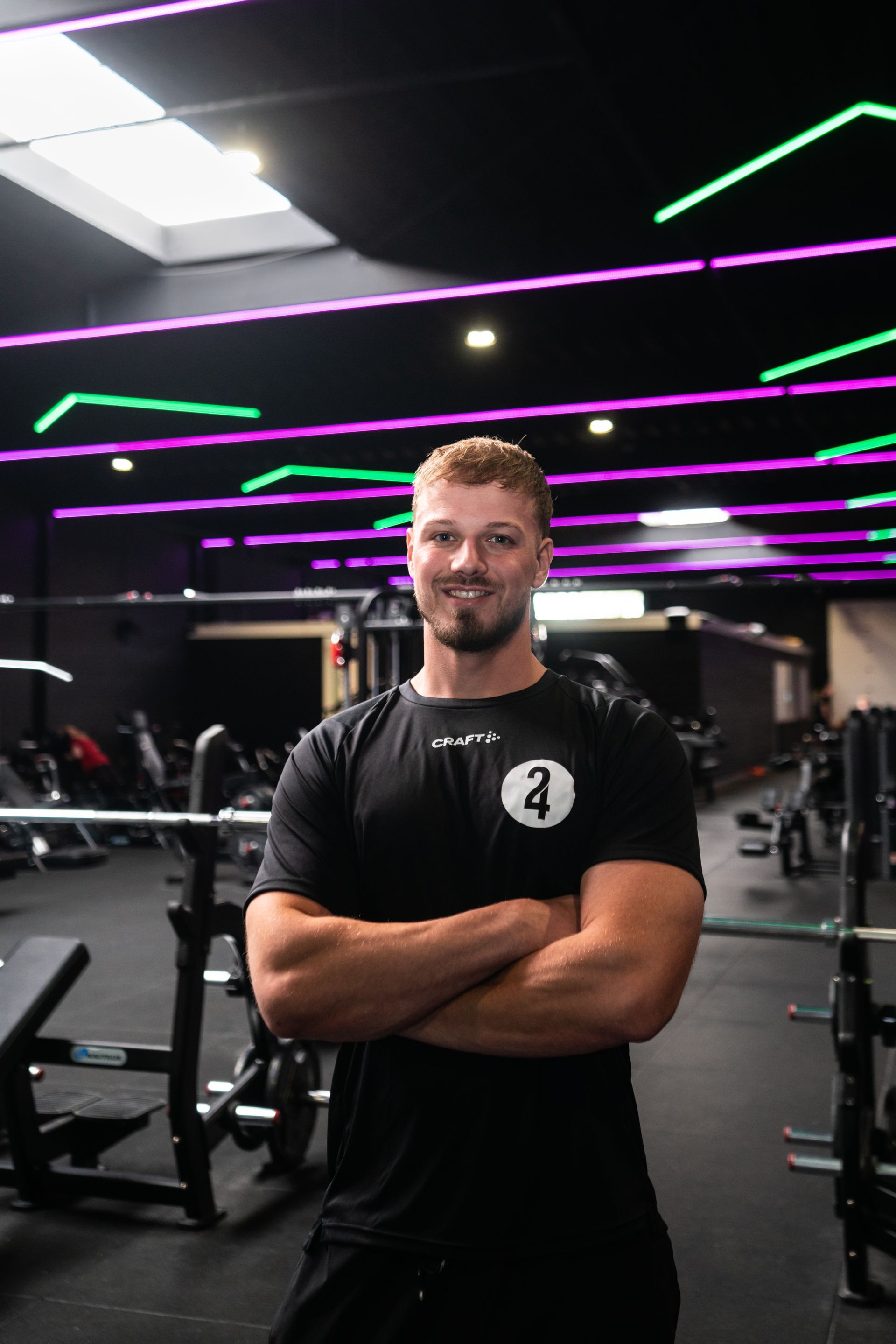 Een man met gekruiste armen lacht in een sportschool. Hij draagt een zwart shirt en op de achtergrond zijn fitnessapparaten te zien.