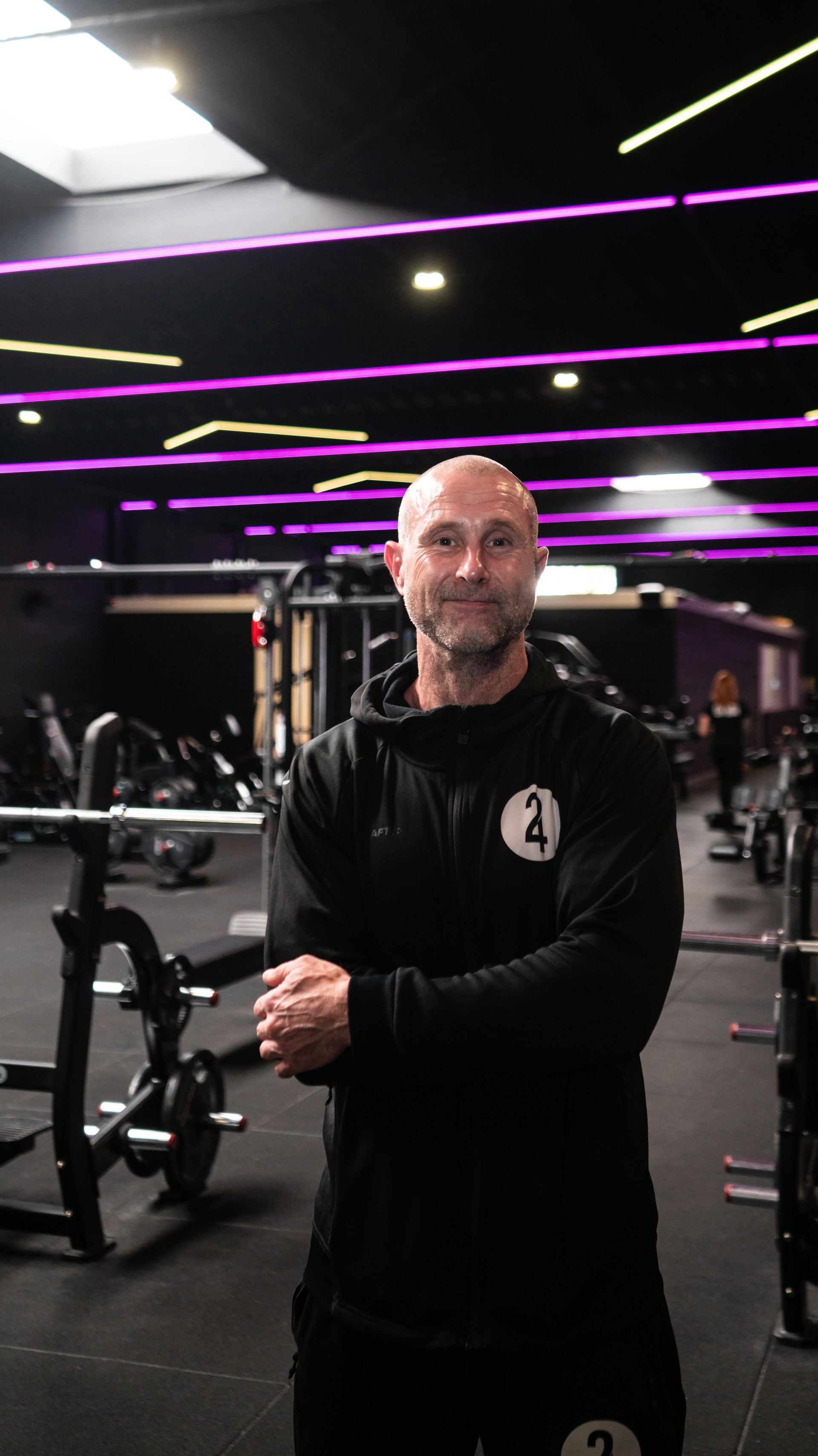 Man met gekruiste armen in een sportschool, omringd door fitnessapparatuur; donker interieur, paarse accenten.