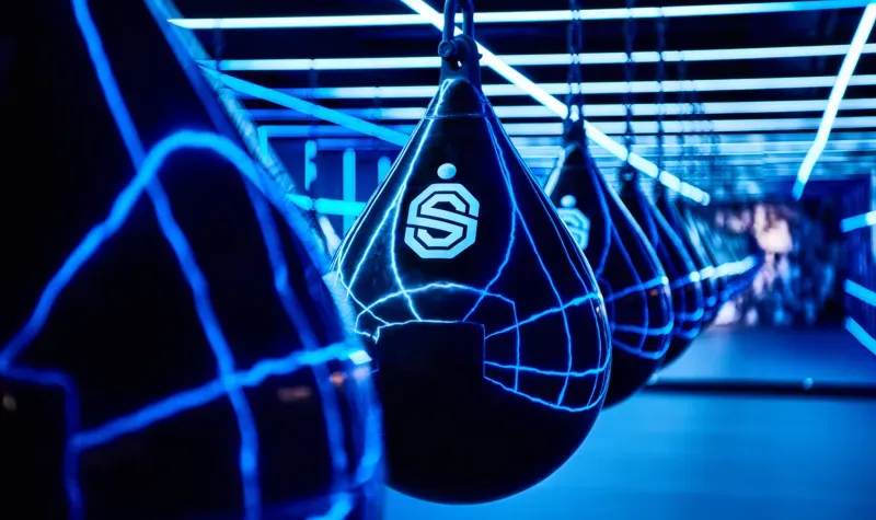 Zwarte, druppelvormige bokszakken hangen in een neonblauw verlichte gymzaal, met op elke zak een logo.