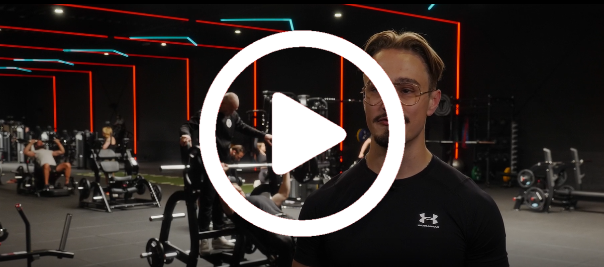 Man in een sportschool; praat in de camera; afspeelknop eroverheen; sportschoolomgeving met apparatuur en mensen op de achtergrond.