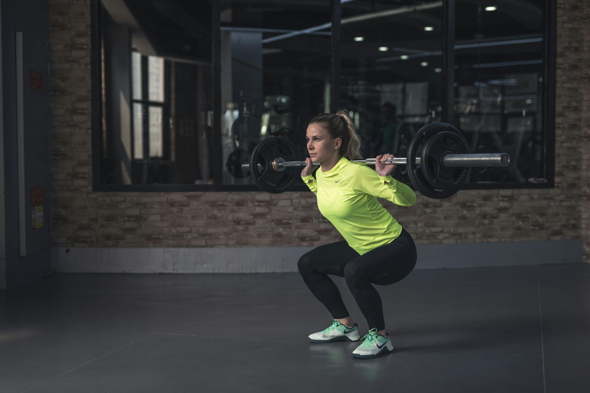 Vrouw in neongroen shirt doet squats met halterstang in de sportschool.