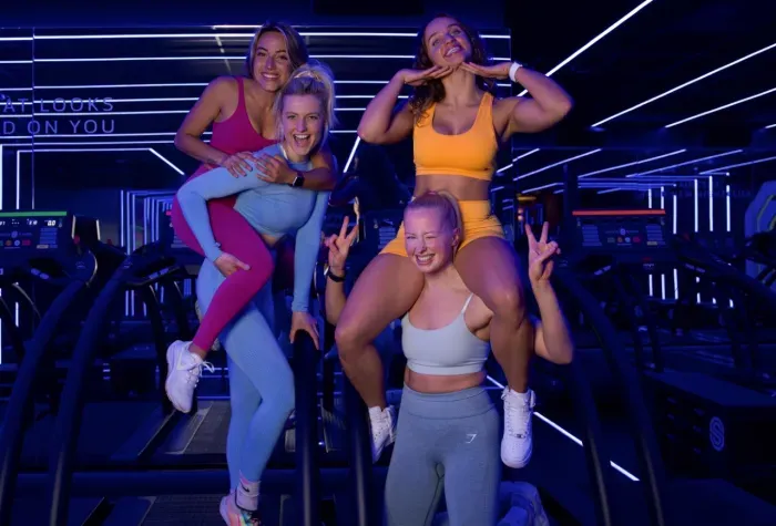 Vier vrouwen in sportkleding poseren op een loopband. Twee rijden lachend op hun rug. De sportschool is blauw verlicht.