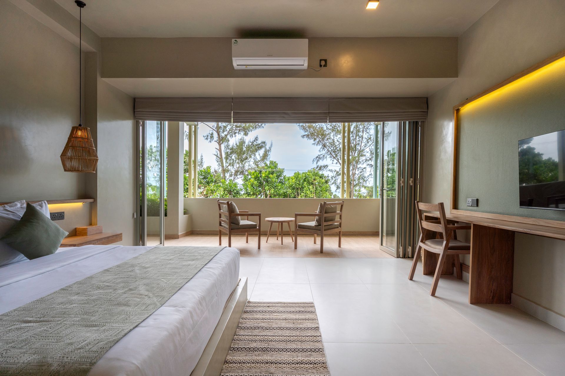 Hotel room with balcony overlooking trees. Light-colored walls, large windows, and a wooden desk.