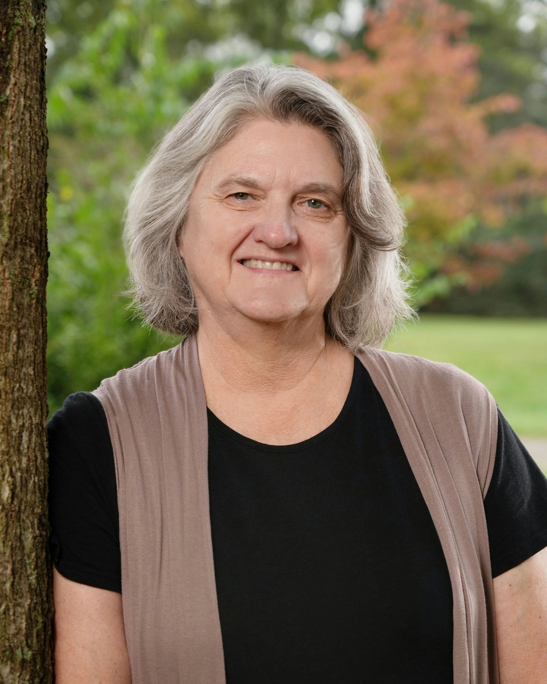 Woman with gray hair, smiling, wearing a black shirt and brown vest, leaning against a tree outside.
