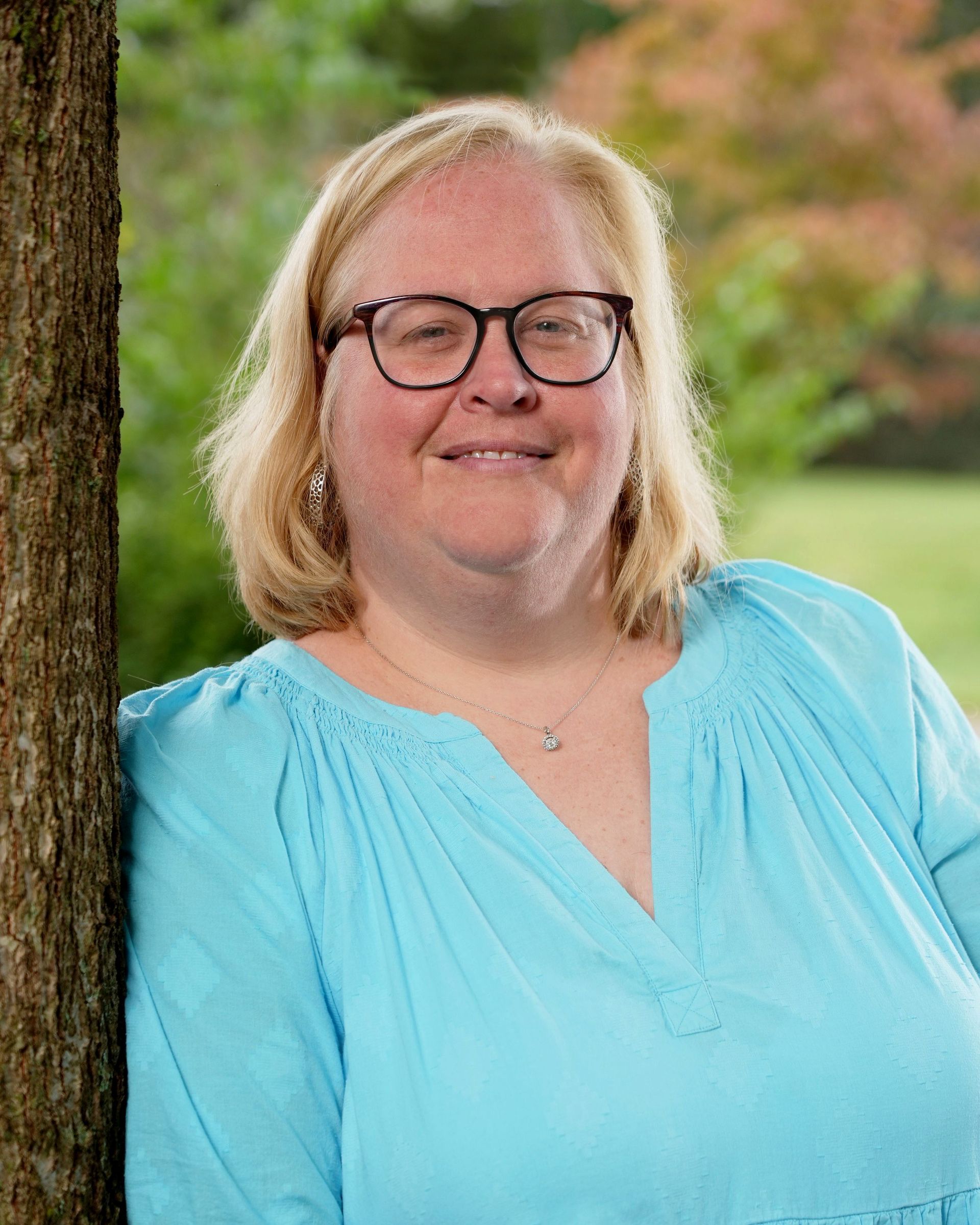 Woman with glasses, light blue shirt, smiles leaning against a tree outdoors.