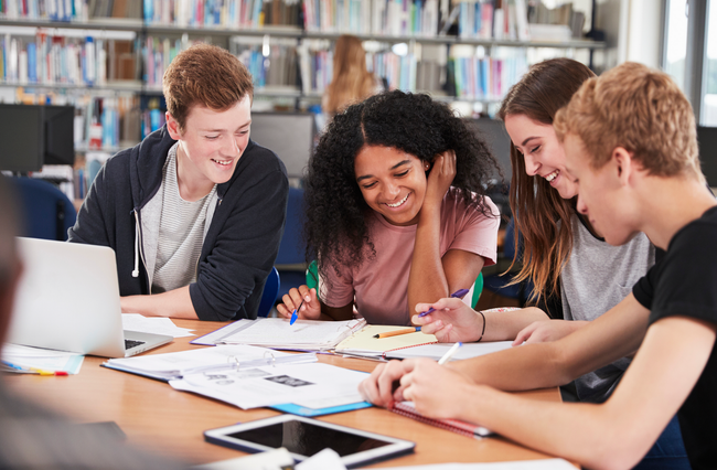 Group of diverse students studying together at a table in a library, smiling and focused.