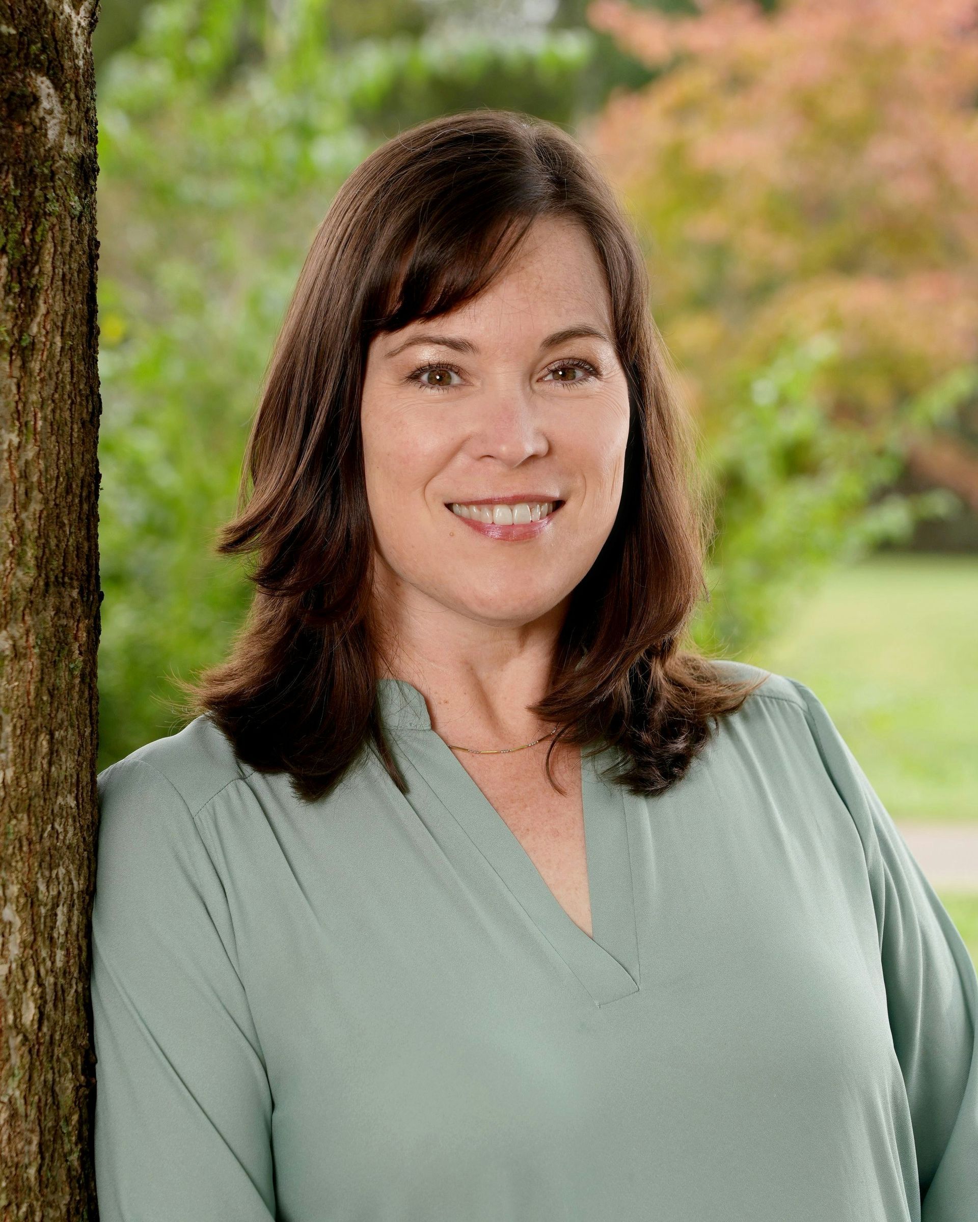 Woman with brown hair, light green shirt, smiling, leaning against a tree in an outdoor park setting.