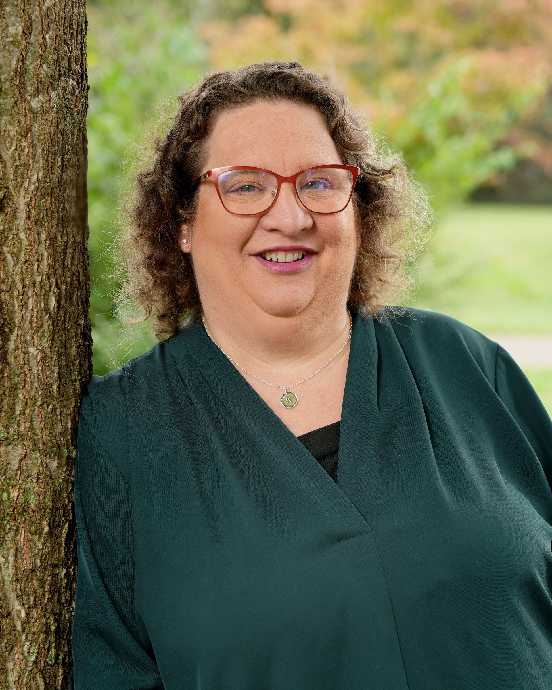 Woman with glasses smiles, leaning against a tree, wearing a green sweater outdoors.